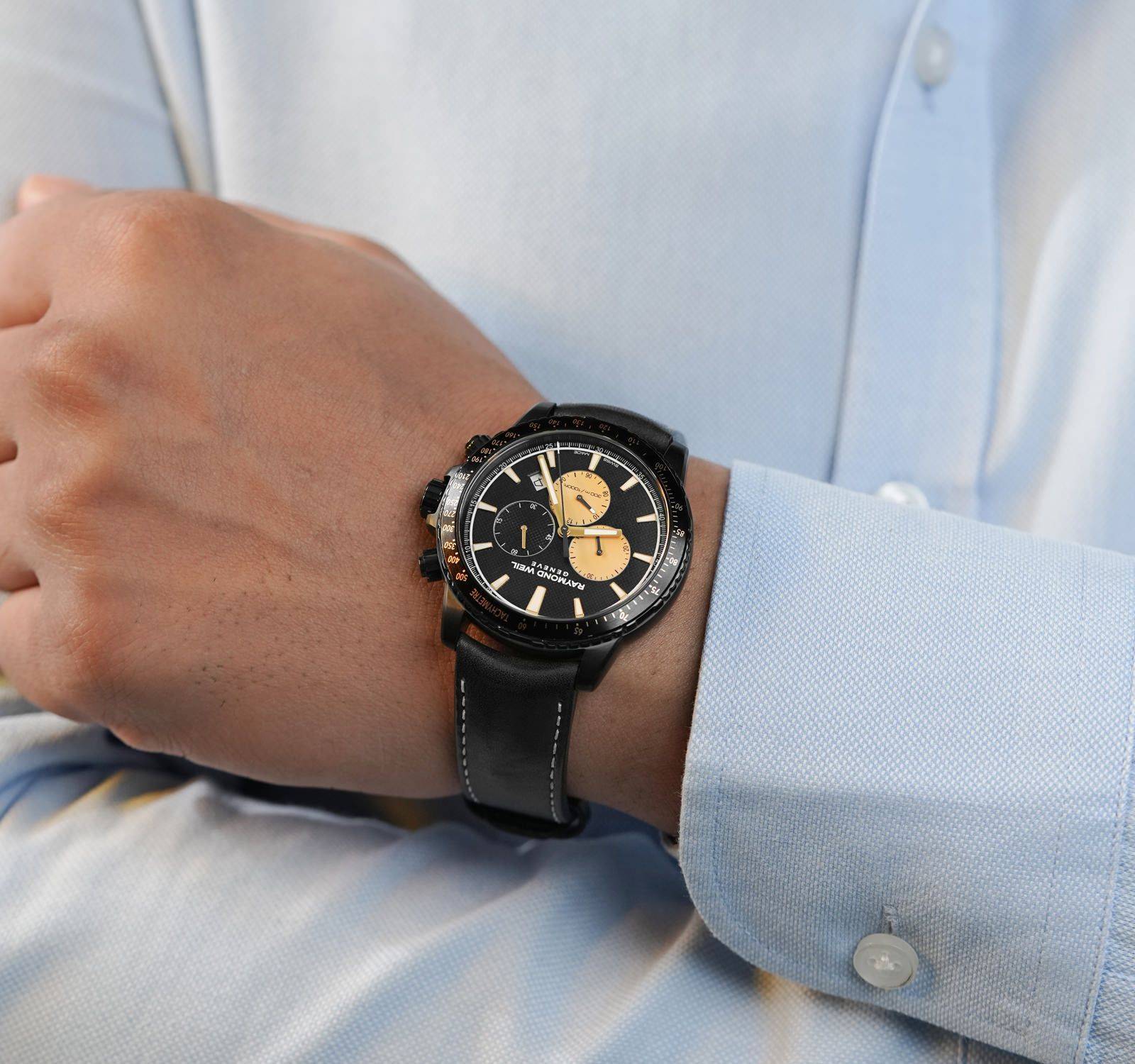 Raymond Weil Tango herenhorloge 43mm met zwarte leren band en goudkleurige chronograafwijzers, gedragen bij een lichtblauw overhemd. Limited Edition model.